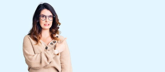 Beautiful young brunette woman wearing casual clothes and glasses pointing aside worried and...