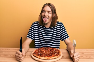 Handsome caucasian man with long hair eating tasty pepperoni pizza winking looking at the camera...
