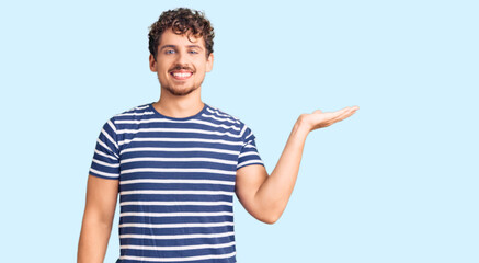 Young handsome man with curly hair wearing casual clothes smiling cheerful presenting and pointing with palm of hand looking at the camera.