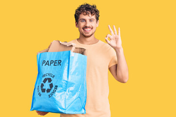 Young handsome man with curly hair holding recycling wastebasket with paper and cardboard doing ok sign with fingers, smiling friendly gesturing excellent symbol