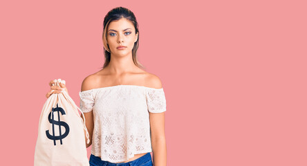 Young beautiful woman holding money bag with dollar symbol thinking attitude and sober expression...