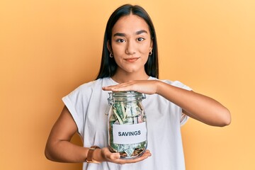 Young asian woman holding jar with savings relaxed with serious expression on face. simple and...