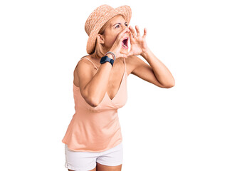 Young blonde woman wearing summer hat shouting angry out loud with hands over mouth