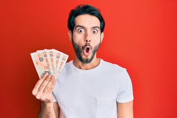 Young hispanic man holding 10 united kingdom pounds banknotes scared and amazed with open mouth for...