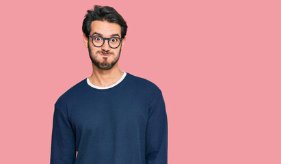 Young hispanic man wearing casual clothes and glasses puffing cheeks with funny face. mouth inflated with air, crazy expression.
