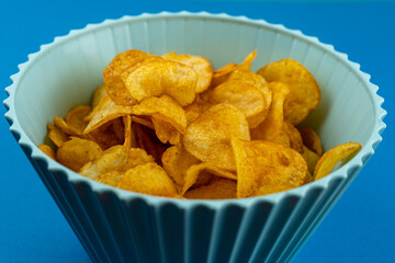 A bowl of potato chips on a blue background with copy space. Party snack.