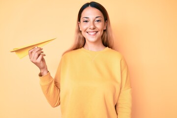 Young brunette woman holding paper airplane looking positive and happy standing and smiling with a confident smile showing teeth
