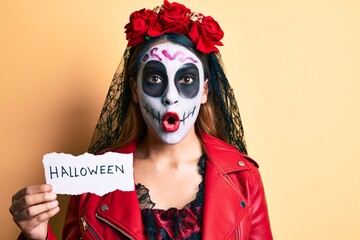 Woman wearing day of the dead costume holding halloween paper scared and amazed with open mouth for surprise, disbelief face