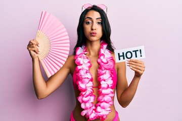 Beautiful hispanic woman wearing bikini and handfan holding hot word puffing cheeks with funny face. mouth inflated with air, catching air.
