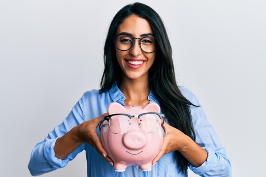 Beautiful hispanic woman holding piggy bank with glasses smiling and laughing hard out loud because funny crazy joke.