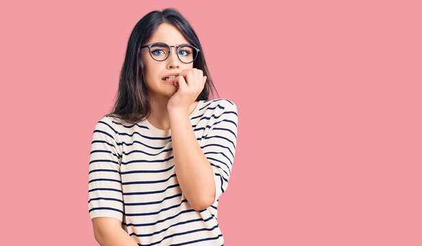 Brunette teenager girl wearing casual clothes and glasses looking stressed and nervous with hands on mouth biting nails. anxiety problem.