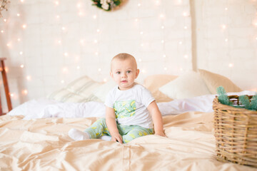 Fototapeta premium Little boy in pajama have fun in in a Scandinavian style bedroom decorated with Christmas garlands and needles on a large bright bed. Christmas mood. Christmas mood. Family time 