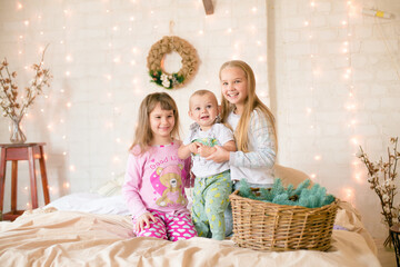 Obraz na płótnie Canvas Cute sisters and little brother in pajamas have fun in in a Scandinavian style bedroom decorated with Christmas garlands and needles on a large bright bed. Christmas mood. Christmas mood. Family time