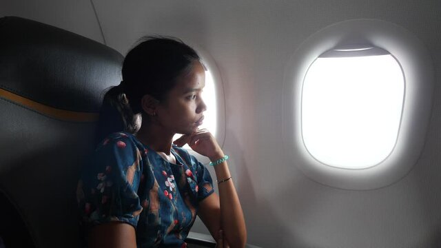 Young Asian woman inside a plane sitting near the window and watching outside.