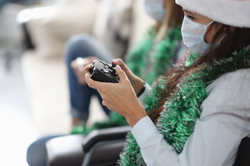 Two girlfriends are sitting on the couch with game joysticks in santa claus hats and protective medical masks. Entertainment for New Year and Christmas in conditions of quarantine and coronavirus