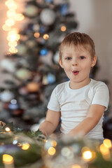 A beautiful boy of 4-5 years old is sitting or standing near the Christmas tree. He dreams of a holiday and gifts, makes a wish. The concept of the winter holidays.