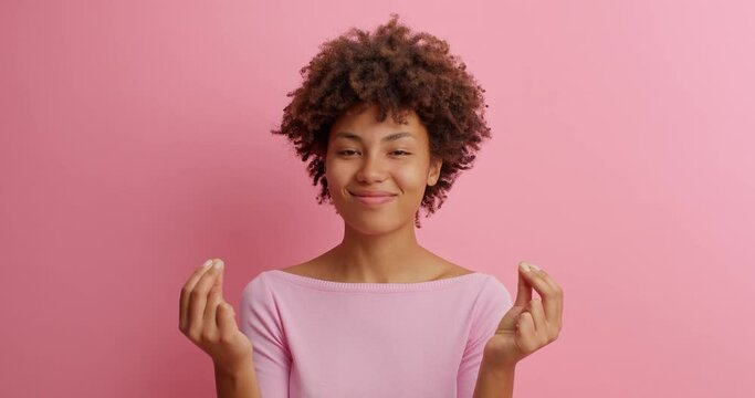 Cunning pleased young Afro American woman makes money gesture has idea of big profit says I know how to earn imitates to scatter cash dressed in casual clothing poses against pink background