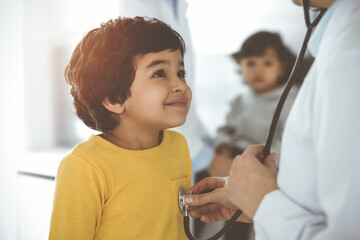 Woman-doctor examining a child patient by stethoscope in sunny clinik. Cute arab boy at physician...