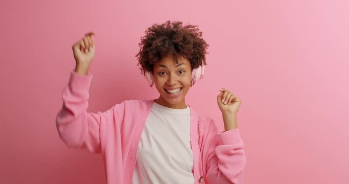 Carefree joyful African American woman moves energetically keeps arms raised feels very glad listens favorite music via headphones poses against rosy background. Feeling rhythm of every tune