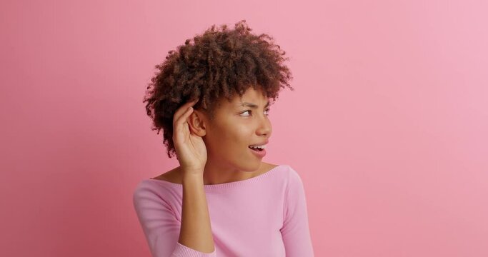 Cute curious woman keeps hand near ear tries to overhear something very interesting listens somebodys secret looks away likes collecting rumors poses against pink background. Eavesdropping concept