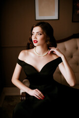 Lovely young woman in black evening dress and pearl necklace, classic make-up in the style of Coco Chanel, on a sofa in a classic interior. Beauty and fashion.