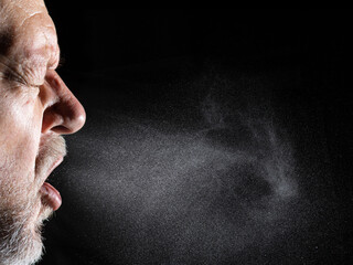 A man sprays aerosols into the air while speaking. The background is black.