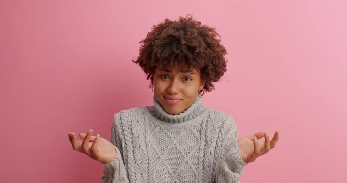 Attractive hesitant dark skinned Afro American woman spreads hands sideways as being clueless and doubtful cannot make choice poses unaware dressed in casual jumper isolated over pink background