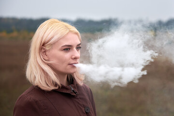 Young blonde woman exhales cigarette smoke on nature.