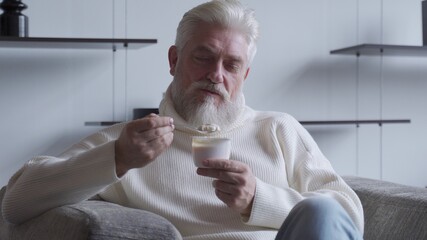 A handsome elderly man with a gray beard eats yogurt holding a spoon close to his mouth