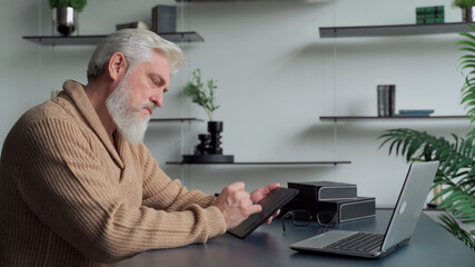 An elderly man with a gray beard draws with a pen on a tablet at home in the living room