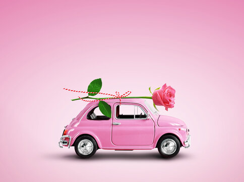 Pink toy car delivering pink rose flower on pink background. Flowers delivery, women day, valentine day.