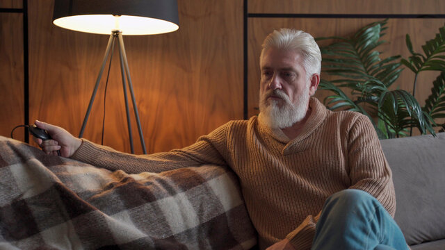 An elderly man with a gray beard sitting on the sofa turns on and off the lamp. A brooding older man is depressed