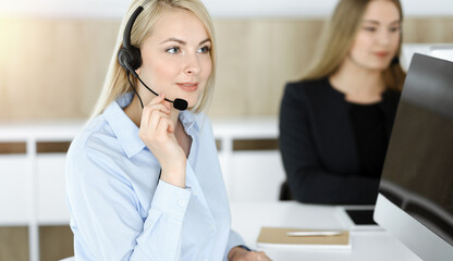 Blonde business woman sitting and communicated by headset in sunny call center office