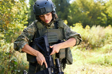 confident soldier woman loading rifle in the forest, alone, looking at gun, wearing military wear