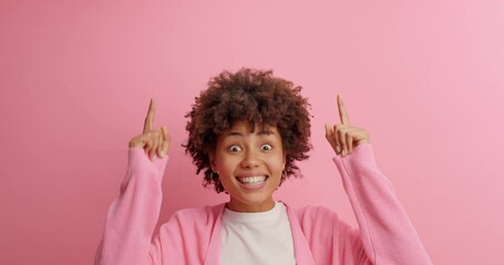 Positive Afro American woman in casual clothing points index fingers up attracts your attention and asks to look shows advertisement with happy smile against rosy background. Promotion concept