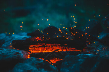 Fototapeta na wymiar Vivid smoldered firewoods burned in fire close-up. Atmospheric background with orange flame of campfire and blue smoke. Warm full frame image of bonfire. Glowing embers in air. Bright sparks in bokeh