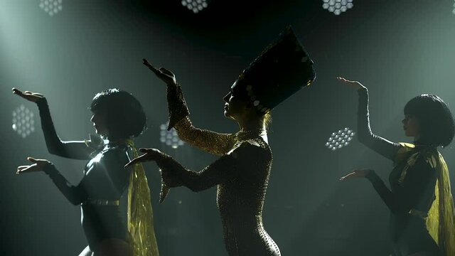 The actress, dressed as the queen of ancient Egypt, dances with two women in a dark studio. Silhouettes of slender women on a smoky background with backlight. Close up.