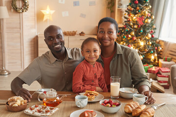 Portrait of happy African-American family enjoying tea and sweets while celebrating Christmas at home in cozy home interior