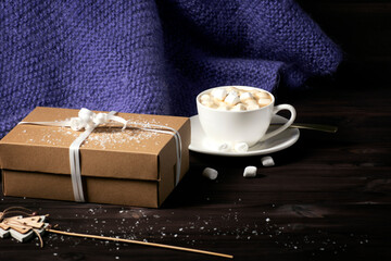 This photo shows a knitted purple blanket, a cup of hot cocoa with marshmallows, and a gift box on a dark wooden background with snow.