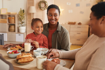 Obraz na płótnie Canvas Portrait of cute African-American girl enjoying breakfast with mom and grandma in cozy home interior