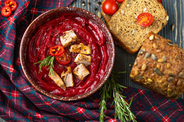 Thick tomato soup with meat slices in a ceramic plate. Next to the plate are a checkered serving napkin, bread, tomatoes, and a sprig of rosemary. The view from the top.
