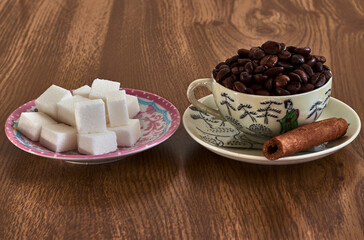 Porcelain cup on a saucer, with roasted coffee beans and a cinnamon stick, next to a plate with square sugar cubes
