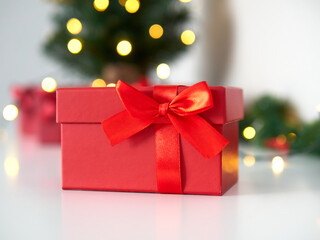 Close up of a red gift box. Beautiful gift box with a red ribbon and a bow. Background with green Christmas tree and lights. Christmas time and gift time