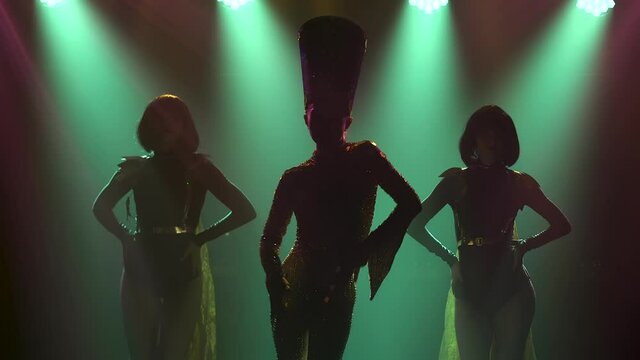 Silhouettes of a young woman in a costume and headdress of Cleopatra or Nefertiti, dancing with two women in a dark studio. Theatrical performance on a smoky background with studio lights. Close up.
