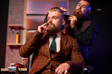 stylish male enjoy haircut at the hairdresser, he is smoking during process of cutting, barber cuts the hair on the client's head while he is smoking cigarette, wearing formal wear