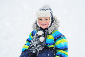 a small child boy in a bright striped jacket sitting on a sledge smiles against the background of snowfall. Winter entertainment