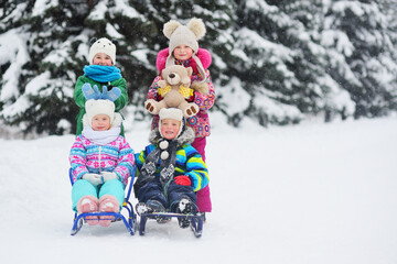 Fototapeta na wymiar a group of children in bright colored winter clothes on a sled with toys in their hands smile and play against the background of snow, snowfall and winter forest