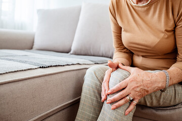 Elderly woman seated on couch touches knee suffers from repeated painful feelings on knee pain...