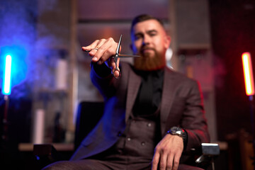 close-up photo of scissors in hands of male barber at work place, he is sitting on chair and...