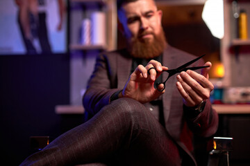 close-up photo of scissors in hands of professional male barber, handsome guy in suit examining, checking scissors for future cutting hair of clients